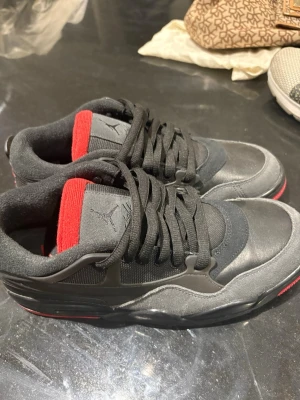 Svarta Air Jordan sneakers med röd detalj - Säljer ett par svarta Air Jordan sneakers med röda detaljer på insidan och sulan. Skorna har lågt skaft, svart mocka och läder, samt klassisk Jumpman-logga på plösen. Snygg och tidlös design som passar till streetwear och casual outfits.