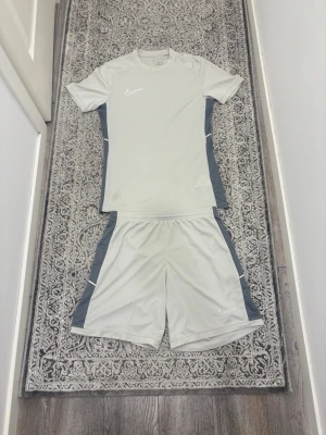 Nike t-shirt och shorts - Snygg vit t-shirt/shorts från Nike med gråa paneler längs sidorna och diskret Nike-logga på bröstet. T-shirten har rund halsringning och korta ärmar, perfekt för sportiga tillfällen. Tillverkad i ett lätt och funktionellt syntetmaterial.