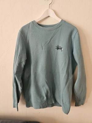 Ljusblå crewneck från Stüssy - Ljusblå sweatshirt från Stüssy med klassisk logga tryckt på bröstet och stor logga på ryggen. Tröjan har rund halsringning, långa ärmar och ribbade muddar. Perfekt för en avslappnad streetwear-look.