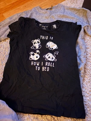 Svart panda-pyjamas med tryck - Mysig svart pyjamas med gulligt pandamotiv och texten 'This is how I roll to bed' på tröjan. Byxorna har matchande pandamönster och dragsko i midjan. Perfekt för chillkvällar hemma. Materialet känns mjukt och bekvämt mot huden.