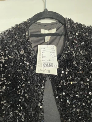 Svart paljettcardigan från H&M - Glittrig svart cardiganjacka från H&M täckt med paljetter. Kort modell med öppen front och korta ärmar, perfekt för att lyfta vilken outfit som helst. Fodrad insida för extra komfort och snygg finish.