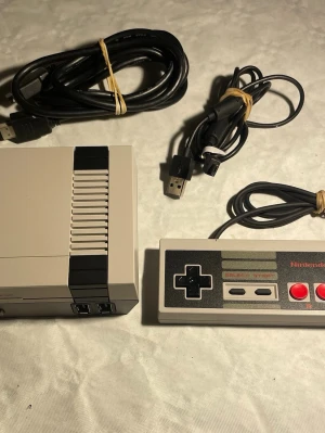 Nintendo Classic Mini NES konsol - Nintendo Classic Mini NES är en kompakt retro-spelkonsol i grå plast med svart detalj, inspirerad av originalet från 80-talet. Medföljer gör en klassisk NES-handkontroll, HDMI-kabel och USB-strömkabel. Perfekt för dig som vill spela gamla favoriter på modern TV.