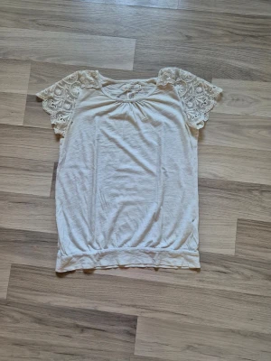 Vit topp med spetsärmar - Söt vit topp med korta ärmar i spets och rund halsringning. Toppen har en avslappnad passform och är gjord i mjukt bomullsmaterial. Perfekt för en fräsch och feminin look. Har 2 hål, se bild, som säkert går att laga för den händiga. 