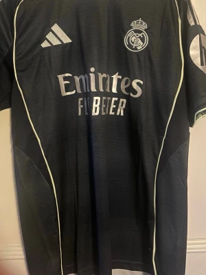 Real Madrid 24/25 Valverde 8 - Snygg svart Real Madrid matchtröja från säsongen 24/25 med klubbmärke och sponsortryck. På ryggen så står det Valverde 8. Tröjan har korta ärmar, rund hals och tunna vita detaljer längs sidorna. Perfekt för fotbollsträning eller att bära som supporter. Materialet är lätt och andas bra.