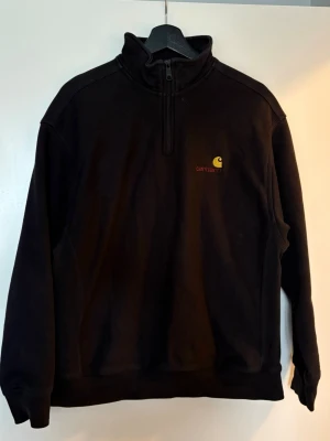 Svart Carhartt halfzip hoodie - Svart hoodie från Carhartt med halv dragkedja och hög krage. Liten gul Carhartt-logga och röd text på bröstet. Mjuk och skön bomullskänsla, perfekt för en avslappnad streetwear-look. Långa ärmar och ribbade muddar.
