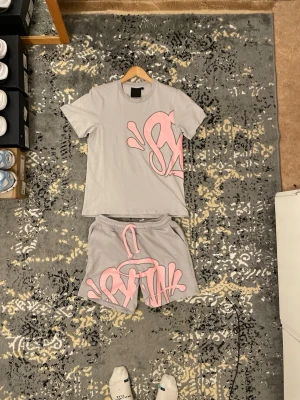 Grått syna world set med rosa tryck - Säljer ett grått set med t-shirt och shorts, båda med stort rosa grafiskt tryck på både fram- och baksidan. Setet har en avslappnad passform och är gjort i mjukt material, perfekt för chill dagar. Märket är ej angivet.