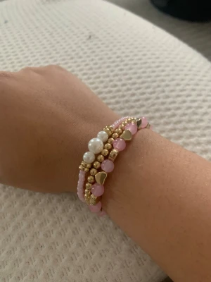 Rosa och pärlarmband med gulddetaljer - Säljer ett snyggt armband med flera rader av rosa och vita pärlor, guldfärgade kulor och små hjärtan. Armbandet har en trendig och ungdomlig look med mixade material och färger som verkligen sticker ut.