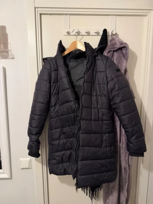 Svart lång dunjacka med huva - Säljer en svart lång dunjacka med huva och quiltad design. Jackan har dragkedja framtill och är perfekt för kalla dagar. Den är stilren och enkel att matcha med olika outfits. Passar dig som vill ha en varm och snygg jacka under höst och vinter.aldrig använt