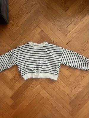 Randig croppad topp från Dazy - Säljer en vit och svart randig croppad topp från Dazy. Toppen har långa ärmar och en bred vit mudd vid nederkanten. Perfekt för dig som gillar en avslappnad och trendig stil. Materialet är mjukt och känns som bomull.