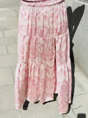 Rosa långkjol - Säljer en jättefin rosa långkjol med ett mjukt och flowy fall. 💕 Har en smockad, stretchig midja och ett tunt, luftigt material som faller väldigt fint. Mönstret och modellen liknar verkligen den virala kjolen från Maje.  I utmärkt skick – inga skavanker eller tecken på användning. Märkt storlek L, men passar XS–M beroende på hur man vill att den ska sitta tack vare den elastiska midjan.  Perfekt till sommaren, semester eller en avslappnad men chic look! ✨