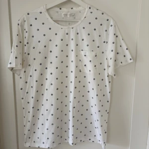 Vit prickig t-shirt med ficka från Emilio - Säljer en vit t-shirt från Emilio med små svarta (eller väldigt mörkblå. Kan ej se skillnaden) pricksr över hela tröjan och en bröstficka. Klassisk rund halsringning och korta ärmar. Tillverkad i mjuk bomull, perfekt för en avslappnad fräsch stil.