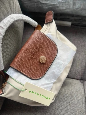 Beige handväska från Longchamp - Beige handväska från Longchamp med bruna läderdetaljer och guldfärgade metalldetaljer. Väskan har ett klassiskt lock med tryckknapp och präglad logga samt ett kort handtag i läder. Axelrem tillkommer och prislappar tillkommer.💕