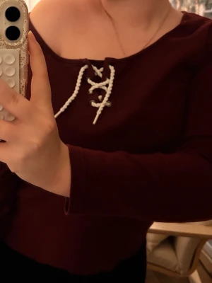 Vinröd topp med snörning - Vinröd långärmad topp med snygg snörning framtill vid halsen. Toppen har en offshoulder-design och är tillverkad i ett mjukt bomullsliknande material. Perfekt för dig som vill ha en trendig men även vintage look.💞