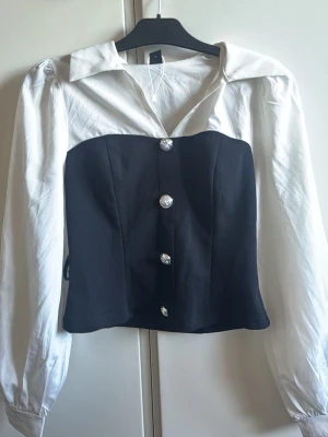 Svartvit blus med markerad midja - Trendig svartvit blus med långärmade vita ärmar och krage, samt svart figursydd front med dekorativa silverknappar. Snygg kontrast mellan färgerna och en modern, uppklädd look. Perfekt för dig som vill sticka ut med stilren design.