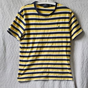 Randig t-shirt från Polo Ralph Lauren - Snygg randig t-shirt från Polo Ralph Lauren i slim fit-modell. T-shirten har gula, vita och marinblå ränder samt en liten broderad logga på bröstet. Tillverkad i mjuk bomull och har korta ärmar och rund hals.