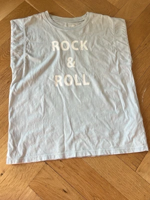 Ljusblå ärmlös topp med tryck - Snygg ljusblå ärmlös topp med texten 'ROCK & ROLL' i vitt framtill. Rak passform och rund halsringning. Perfekt för dig som gillar en avslappnad och cool stil. Tillverkad i mjuk bomull för skön känsla hela dagen.