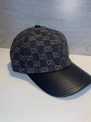 Svart keps från Gucci med GG-mönster - Säljer en svart keps från Gucci med klassiskt GG-monogram i grått över hela tyget. Skärmen är i svart läderimitation som ger en lyxig känsla. Perfekt accessoar för dig som vill ha en trendig och exklusiv look.