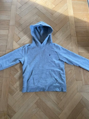 Grå hoodie från Ralph Lauren - Snygg grå hoodie från Ralph Lauren med klassisk broderad logga på bröstet. Hoodien har en stor huva och en praktisk magficka. Tillverkad i mjukt bomullsmaterial som är skönt att ha på sig. Perfekt för en avslappnad och stilren look.