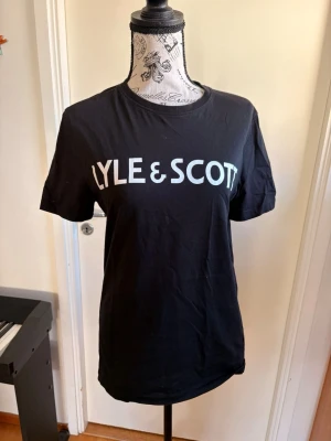 Svart Lyle & Scott t-shirt - Snygg svart t-shirt från Lyle & Scott med stor vit logga över bröstet. Klassisk rund halsringning och korta ärmar. Perfekt till jeans eller shorts för en clean och enkel stil. Passar till 14-15 år