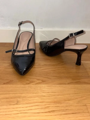Svarta kittenheels - Snygga svarta pumps i lack med spetsig tå och smala remmar med silverspännen. Skorna har öppen häl och medelhög klack som ger en elegant siluett. Perfekta för dig som gillar stilrena och trendiga skor.