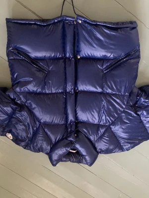 Mörkblå dunjacka från Moncler - Snygg mörkblå dunjacka från Moncler med glansig finish och två snedställda bröstfickor med dragkedja. Jackan har hög krage och klassisk Moncler-logga på ärmen. Tillverkad i Frankrike och fylld med äkta dun för maximal värme.