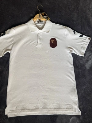 Vit polo t-shirt från A Bathing Ape - Vit polo t-shirt från A Bathing Ape med klassisk krage och knappslå. Bröstet pryds av det ikoniska BAPE-apans logomärke i brunt. Svarta detaljer på ärmarna och camo-tag vid kragen. Perfekt för en clean och stilren look.