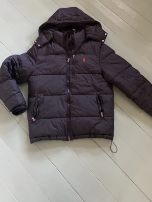 Svart dunjacka från Polo Ralph Lauren - Snygg svart dunjacka från Polo Ralph Lauren med huva och dragkedja. Jackan har två fickor med dragkedja och en röd broderad logga på bröstet. Perfekt för kalla vårdagar och riktigt skön att bära. Klassisk och stilren design.