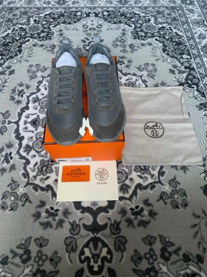 Grå H E R M E S sneakers med vit sula - Snygga grå sneakers  med mesh och mockadetaljer. Skorna har en vit och grå sula med stort H-mönster undertill och klassisk snörning. Kommer med originalkartong och dustbag. Perfekta för dig som gillar exklusiva och stilrena sneakers.