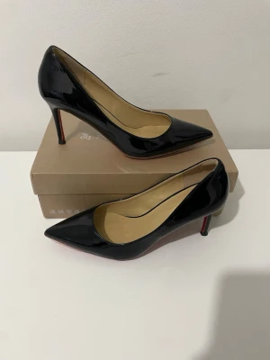 Svarta lackade pumps med smal klack - Snygga svarta pumps i lackat material med spetsig tå och smal hög klack. Insidan är beige och sulan har en röd ton. Perfekta för att lyfta vilken outfit som helst med en elegant touch.