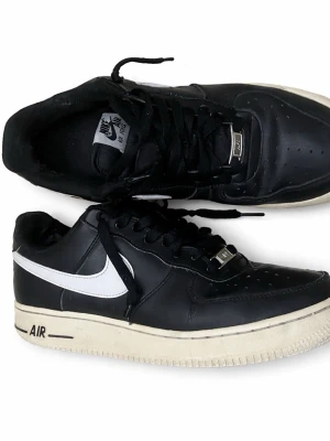 Nike Air Force 1 svart/vit sneaker - Snygga Nike Air Force 1 sneakers i svart läder med vit swoosh och vit sula. Klassisk låg modell med perforerad tå, svarta skosnören och AIR-detalj på sidan. Perfekt för dig som gillar streetstyle och ikoniska sneakers.
