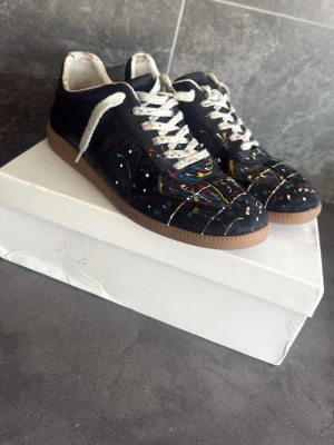 Svarta sneakers med färgstänk - Ett par svarta Maison margiela skor med färg stänkar, mycket bra skick 7,5/10, går för 3000 mitt pris ändast 1300, skriv om ni har frågor om skorna eller om ni vill att jag ska skicka mer bilder!