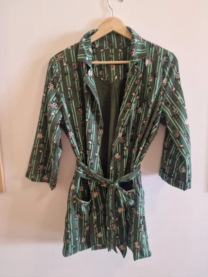 Grön blommig morgonrock med knytskärp - Snygg tunn grön kavaj med blommigt mönster och vertikala ränder. Modellen har trekvartsärmar, klassisk krage och knytskärp i midjan. Materialet känns mjukt och lätt mot huden.