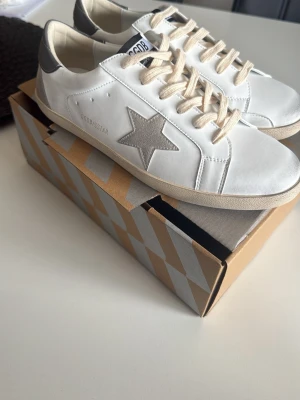 Vita Golden Goose sneakers - Snygga vita sneakers från Golden Goose med grå stjärndetalj på sidan och grå hälkappa. Skorna har beige snörning och klassisk låg siluett. Tillverkade i läder med en stilren och trendig look som passar perfekt till det mesta