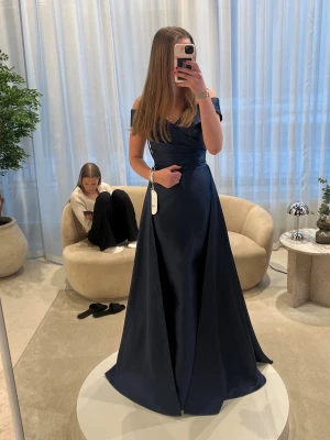 Marinblå balklänning offshoulder - Elegant marinblå långklänning med offshoulder-design och omlott-detalj upptill. Klänningen har en figurnära passform upptill och faller snyggt ner mot golvet. Perfekt för speciella tillfällen och har en lyxig känsla tack vare det glansiga materialet. Den är helt oanvänd och jag säljer den för att jag tvekar på storleken. Nu är den uppe för intressekoll så skriv gärna om ni är intresserade. Prislappen är fortfarande på och jag har aldrig använt den. Säljer pga förfallodatum för returnera har vart