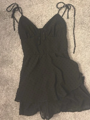 Svart kort jumpsuit från newyorker - Svart kort jumpsuit från newyorker med tunna axelband som knyts, v-ringning och små prickar i tyget. 