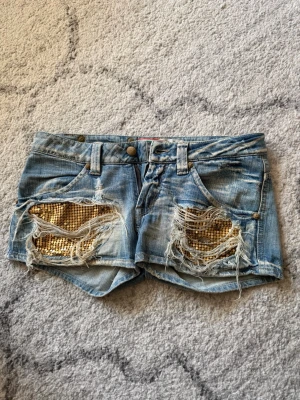Jeansshorts med paljettdetaljer – låg midja - Säljer dessa supersnygga jeansshorts från MET eftersom de tyvärr är för små för mig 💔 De är typ grumligt ljusblå, slitningar framtill med guldiga paljetter under. OBS! Några paljetter är borta men inget man tänker på! Det är låg midja och kort modell som sitter tight. Made in Italy (MET) och känns väldigt välgjorda. Perfekta till sommaren, fester eller semester. Använda men i fint skick, slitningarna är en del av designen.    Hör av dig vid frågor eller om du vill ha fler bilder! 