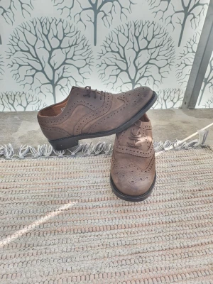 Bruna klassiska loafers i läder - Säljer ett par bruna loafers med snygga broguemönster och klassisk design. Skorna är gjorda i läder och har en låg klack samt rund tå. Perfekta för dig som gillar stilrena och tidlösa skor med detaljer. Passar till många olika outfits. 