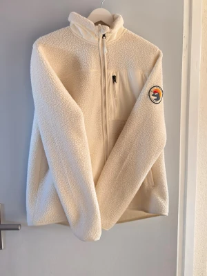 Everest Vit fleece jacka  - Mysig vit teddyjacka med dragkedja framtill, hög krage och en bröstficka med dragkedja. Jackan har en rund patch med färgglad logga på ena ärmen och är tillverkad i fluffigt teddy-material. Perfekt för kalla dagar när du vill vara både varm och snygg. !! Använd 2gngr och helg som ny!!