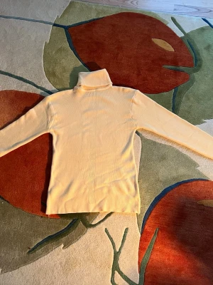 Gul polotröja med diskret mönster - Snygg gul stickad polotröja med lång ärm och ett diskret vertikalt mönster längs mitten. Tröjan har en klassisk polokrage och är tillverkad i ett mjukt, stretchigt material som sitter skönt på kroppen. Perfekt för lager-på-lager under kyliga dagar.