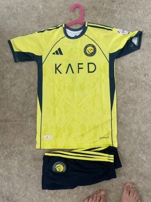Al Nassr Ronaldo matchställ gul/blå - Säljer ett gult och mörkblått matchställ från Al Nassr med Ronaldo 7 på ryggen. Tröjan har klubbmärke, KAFD-logga och Adidas-detaljer. Shortsen är mörkblå med gula ränder och klubbmärke. Perfekt för fotbollsträning eller att bära som supporter.