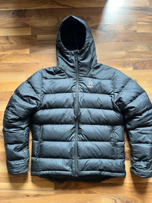Svart dunjacka från Haglöfs med huva - Säljer en svart dunjacka från Haglöfs med huva och dragkedja framtill. Jackan har en puffig design med breda kanaler och diskret logga på bröstet. Perfekt för kalla vinterdagar och riktigt varm tack vare dunfyllningen.