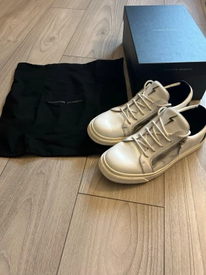 Vita Giuseppe Zanotti sneakers - Snygga vita sneakers från Giuseppe Zanotti i läder med silverfärgade dragkedjor på sidorna och klassisk snörning. Skorna har använts en del men är i ett väldigt bra skick där inga defekter förekommer. Vid fundering är det bara att skriva!