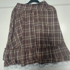 Brun rutig kjol med spetskant - Supersöt brun rutig kjol med volang och vit spetskant längst ner. Kjolen har dragkedja i sidan och är i ett mjukt, lite tjockare tyg som passar perfekt till höst och vinter. Den har en klassisk A-linjeform och fina detaljer som ger en vintagevibe.