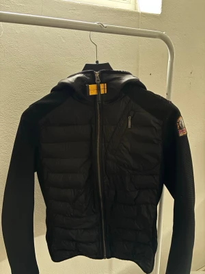 Svart jacka från Parajumpers med huva - Svart jacka från Parajumpers med huva och quiltade partier framtill. Jackan har dragkedja, en bröstficka med dragkedja och ett märke på ärmen. Perfekt för kyligare dagar och har en sportig look.