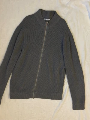 Grå Stickad ziptröja  - Snygg grå ribbstickad ziptröja från Zara med hög krage och dragkedja framtill. Tröjan har långa ärmar och en rak passform som passar perfekt till jeans eller kjol. Materialet är mjukt och ger en avslappnad men stilren look.