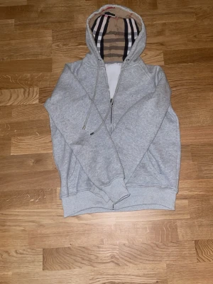 Grå hoodie från Burberry med dragkedja - Snygg grå hoodie från Burberry med klassiskt rutigt foder i huvan och dragkedja framtill. Hoodien har ribbade muddar, dragsko i huvan och diskret Burberry-logga på bröstet. Perfekt för en stilren och avslappnad look. Storlek xs