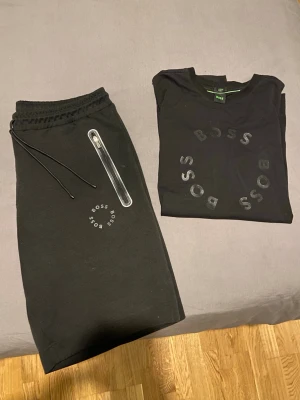 Svart BOSS set med logga - Snyggt svart set från BOSS med matchande t-shirt och shorts. Båda plaggen har BOSS-logga i svart tryck. Shortsen har dragsko i midjan och en ficka med dragkedja. Perfekt för en avslappnad och stilren look.