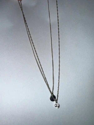 Dubbelhalsband i silverfärg - Säljer ett snyggt dubbelhalsband i silverfärg med två tunna kedjor. Ena hänget är en rund platta och det andra är en liten sten. Perfekt för dig som gillar stilrena accessoarer med en twist.