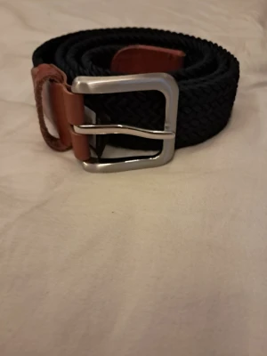 Svart flätat bälte med läderdetaljer - Snyggt svart flätat bälte med bruna läderdetaljer och silvrigt spänne. Perfekt för att lyfta vilken outfit som helst och passar till både jeans och chinos. Enkel att justera tack vare det flexibla materialet.