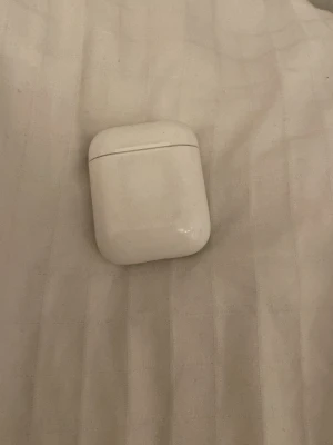 Apple AirPods med laddningsetui - Säljer ett par vita Apple AirPods med det klassiska laddningsetuiet. Trådlösa hörlurar i stilren och kompakt design, perfekt för musik, samtal och poddar. Etuiet är gjort i blank plast och hörlurarna har en smidig passform som sitter bra i öronen.